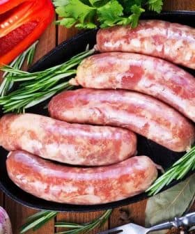Five raw sausages arranged on a black platter with herbs and garlic nearby. - Olive Oil Times