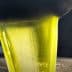 Stream of green olive oil being poured from a metal container into a stainless steel surface. - Olive Oil Times