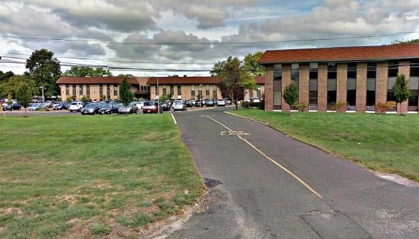 Two commercial buildings with a parking lot in front, surrounded by grass and trees. - Olive Oil Times