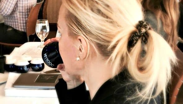 A woman with blonde hair tied in a ponytail holding a blue cup while seated at a table. - Olive Oil Times