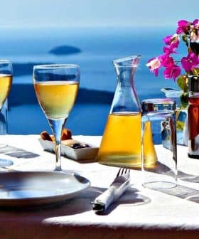A table set with two glasses of wine, a carafe, and a small vase of flowers against a blue ocean background. - Olive Oil Times
