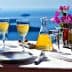 A table set with two glasses of wine, a carafe, and a small vase of flowers against a blue ocean background. - Olive Oil Times