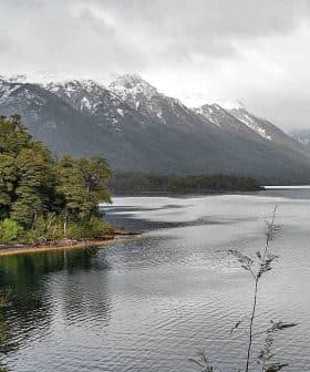 A serene view of a lake surrounded by mountains and dense forest under a cloudy sky. - Olive Oil Times