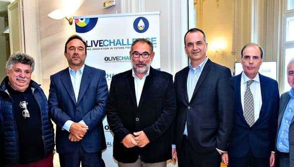 A group of seven individuals posing for a photo at an olive oil event with a banner in the background. - Olive Oil Times
