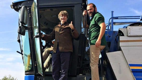 Two men standing on an olive harvesting machine, one giving a thumbs up while the other stands beside him. - Olive Oil Times