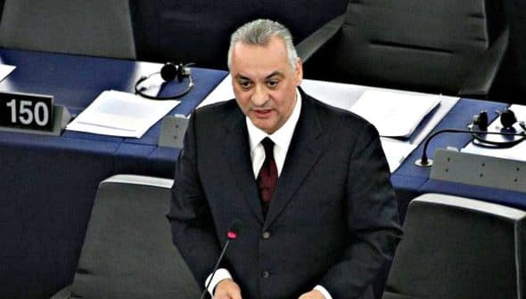 A man speaking at a podium in a legislative assembly with numbered seats visible in the background. - Olive Oil Times