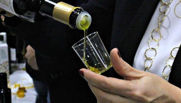 A person pouring olive oil from a bottle into a small glass cup during a tasting event. - Olive Oil Times