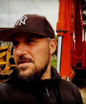 Man wearing a cap standing near an orange excavator on a construction site. - Olive Oil Times