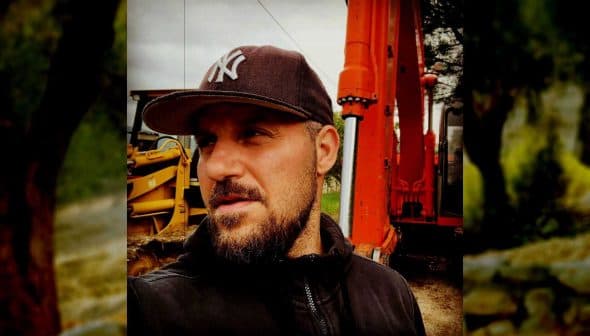 Man wearing a cap standing near an orange excavator on a construction site. - Olive Oil Times