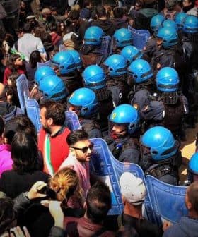 Crowd of people gathered at a demonstration with police officers in riot gear and a police vehicle nearby. - Olive Oil Times