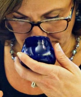 Woman with glasses smelling olive oil from a blue tasting cup while holding a small container. - Olive Oil Times