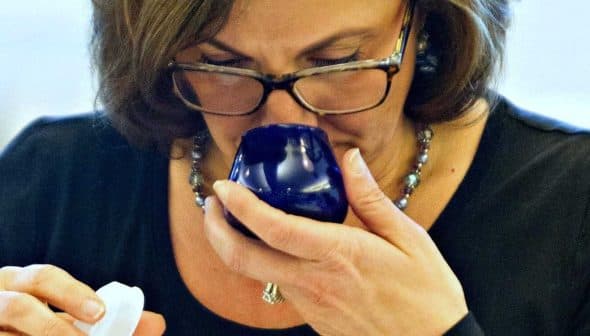 Woman with glasses smelling olive oil from a blue tasting cup while holding a small container. - Olive Oil Times