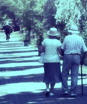An elderly couple walking together on a tree-lined path, both wearing hats and holding hands. - Olive Oil Times