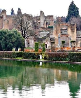 Ancient ruins reflected in a calm water body surrounded by greenery and trees. - Olive Oil Times