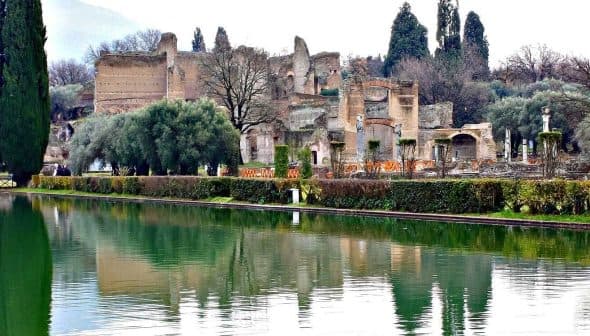 Ancient ruins reflected in a calm water body surrounded by greenery and trees. - Olive Oil Times