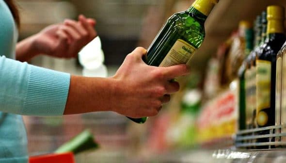A person holding a green bottle of olive oil while shopping in a grocery store aisle. - Olive Oil Times