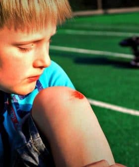 Young boy sitting on a green field with a scraped knee, looking down at the injury. - Olive Oil Times