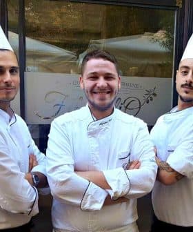 Three chefs wearing white uniforms and hats standing in front of a restaurant entrance. - Olive Oil Times