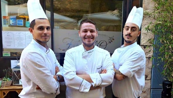 Three chefs wearing white uniforms and hats standing in front of a restaurant entrance. - Olive Oil Times