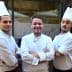 Three chefs wearing white uniforms and hats standing in front of a restaurant entrance. - Olive Oil Times