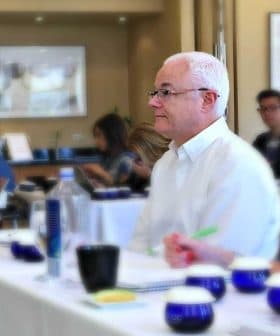 Man with white hair seated at a conference table with participants in the background during a meeting. - Olive Oil Times