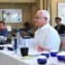 Man with white hair seated at a conference table with participants in the background during a meeting. - Olive Oil Times