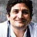 Smiling chef with curly hair wearing a white chef jacket and a gray apron. - Olive Oil Times