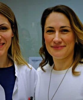 Two women smiling in a laboratory environment, one wearing a lab coat and the other in a white sweater. - Olive Oil Times