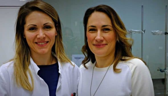 Two women smiling in a laboratory environment, one wearing a lab coat and the other in a white sweater. - Olive Oil Times