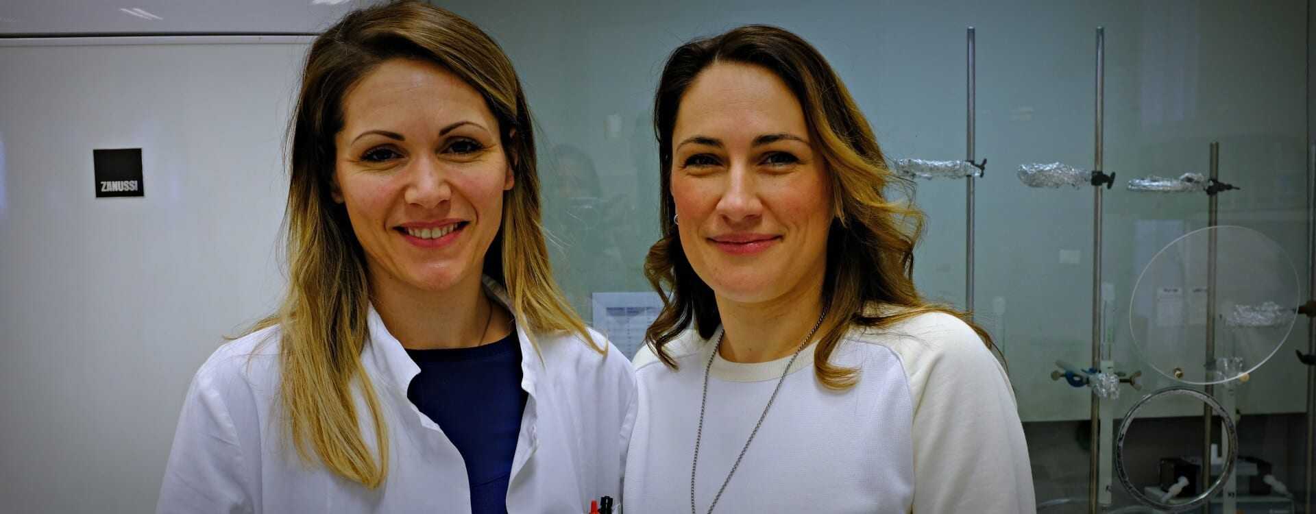 Two women smiling in a laboratory environment, one wearing a lab coat and the other in a white sweater. - Olive Oil Times