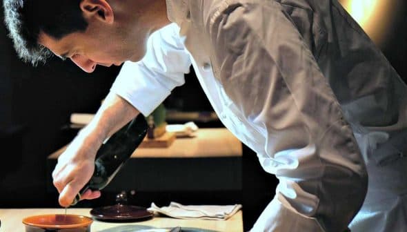 Chef in a white uniform carefully plating a dish with a sauce from a bottle. - Olive Oil Times