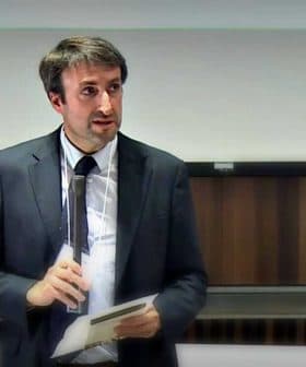 A man in a suit holding a notepad while speaking in front of an audience. - Olive Oil Times