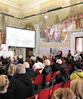 A large audience seated in a historical hall with elaborate murals on the walls. - Olive Oil Times