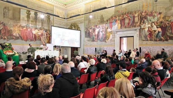 A large audience seated in a historical hall with elaborate murals on the walls. - Olive Oil Times