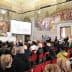 A large audience seated in a historical hall with elaborate murals on the walls. - Olive Oil Times
