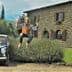A man in a hat and vest jumping joyfully near a vintage car parked by a stone house. - Olive Oil Times