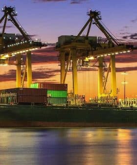 Cargo ship docked at a port with cranes and containers during sunset with colorful sky. - Olive Oil Times