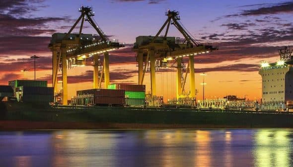 Cargo ship docked at a port with cranes and containers during sunset with colorful sky. - Olive Oil Times