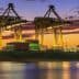 Cargo ship docked at a port with cranes and containers during sunset with colorful sky. - Olive Oil Times