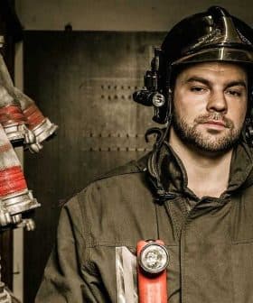 Firefighter wearing a helmet and uniform, standing in a storage area with hoses and equipment in the background. - Olive Oil Times