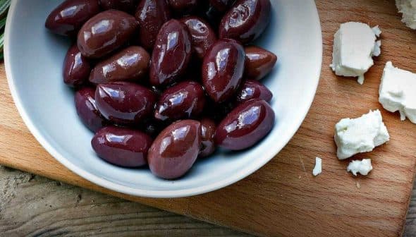 Bowl of Kalamata olives next to feta cheese on a wooden cutting board. - Olive Oil Times