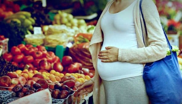 Pregnant woman standing in front of a fruit market display with various fruits. - Olive Oil Times