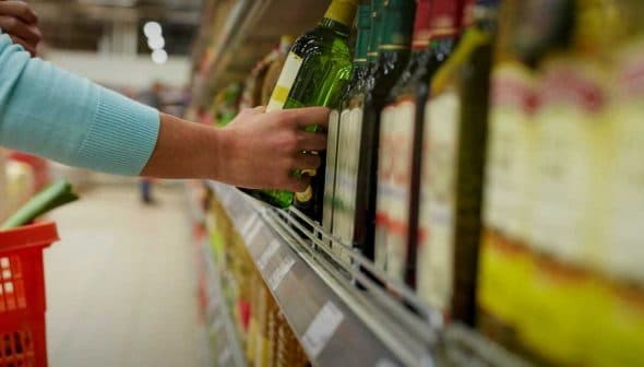 A person reaching for a bottle of olive oil on a supermarket shelf. - Olive Oil Times