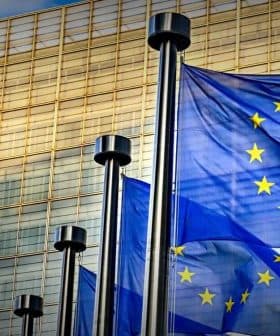 Three European Union flags waving in front of a modern glass building. - Olive Oil Times