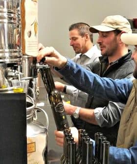 Three men filling bottles with olive oil from a large container in a processing facility. - Olive Oil Times