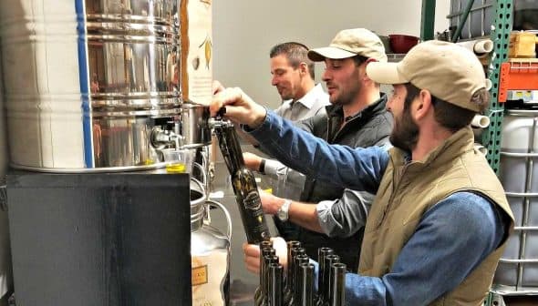 Three men filling bottles with olive oil from a large container in a processing facility. - Olive Oil Times