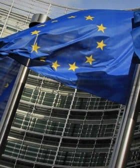 Multiple European Union flags waving in front of a modern glass building. - Olive Oil Times
