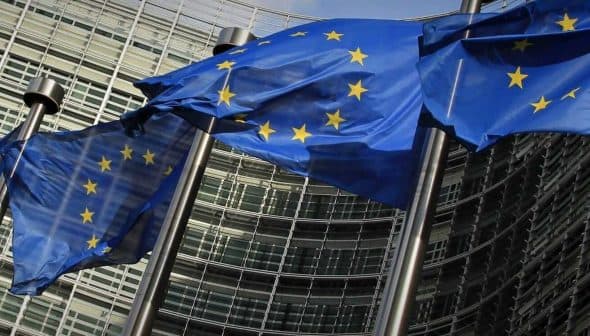Multiple European Union flags waving in front of a modern glass building. - Olive Oil Times