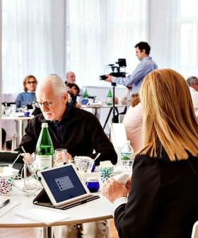 A group of individuals seated at tables during a meeting, with a photographer capturing the moment. - Olive Oil Times