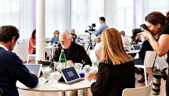 A group of individuals seated at tables during a meeting, with a photographer capturing the moment. - Olive Oil Times